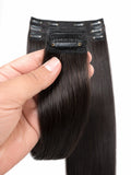 A hand holding a bundle of straight, dark brown to near-black clip-in hair extensions against a plain white background.