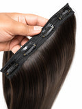 A close-up of a hand holding a clip-in hair extension with straight, dark brown hair and black elastic bands with metal clips, shown against a white background.